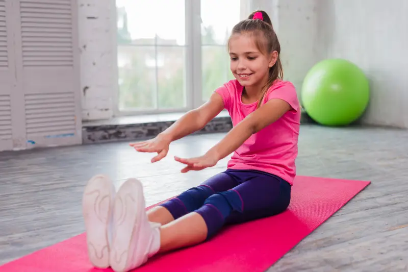 A child in a pink shirt and purple leggings sits on a yoga mat, reaching forward during an exercise session, with a green exercise ball nearby